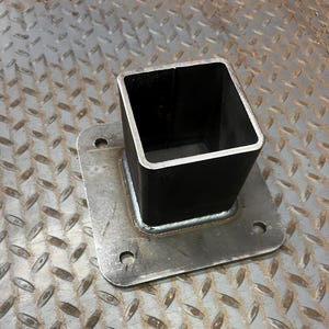 May include: A black, square metal tube welded to a square metal plate with four holes. The metal has a silver color and a diamond plate pattern in the background. The tube is open at the top and appears to be hollow.