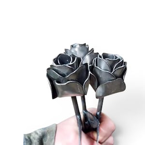 May include: Three metal roses with dark gray petals and stems. The handcrafted flowers have a textured appearance and are held together. The roses are a unique decorative item.