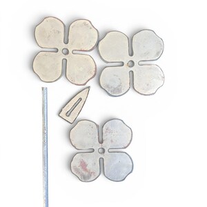 May include: An overhead shot shows four metal flower-shaped objects with a distressed finish, along with a small metal bar and a metal hair clip. The flowers have four petals and a hole in the center. The objects are on a white background.