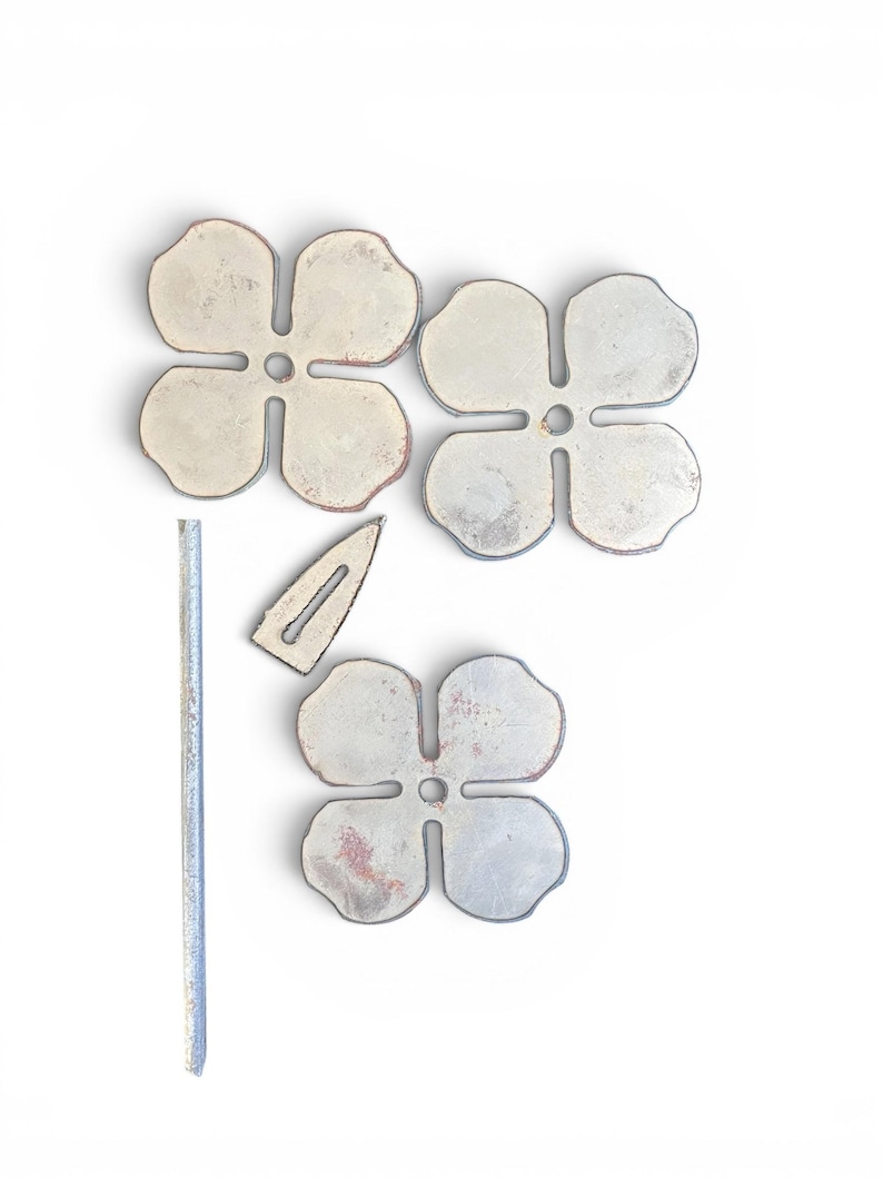 May include: An overhead shot shows four metal flower-shaped objects with a distressed finish, along with a small metal bar and a metal hair clip. The flowers have four petals and a hole in the center. The objects are on a white background.