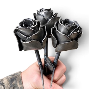 May include: A bouquet of three handcrafted metal roses featuring dark gray, burnished steel petals with visible welded seams. Each rose is attached to a slender, rigid metal stem, showcasing a raw industrial finish with metallic textures.