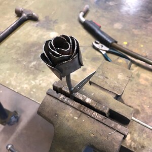 May include: A handcrafted metal rose sculpture, featuring detailed petals and a stem, is held in a vise. The rose is made of dark metal and appears to be welded. Tools like a hammer and welding equipment are visible in the background.