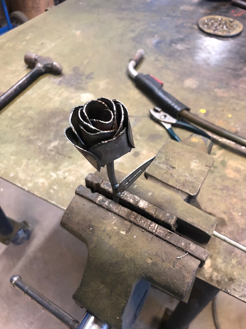 May include: A handcrafted metal rose sculpture, featuring detailed petals and a stem, is held in a vise. The rose is made of dark metal and appears to be welded. Tools like a hammer and welding equipment are visible in the background.