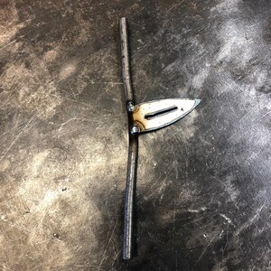 May include: A handcrafted metal tool with a blade-like shape and a vertical rod. The blade is attached to the rod with small rivets. The metal has a silver color and appears to be welded. The tool is likely used for metalworking or a similar craft.