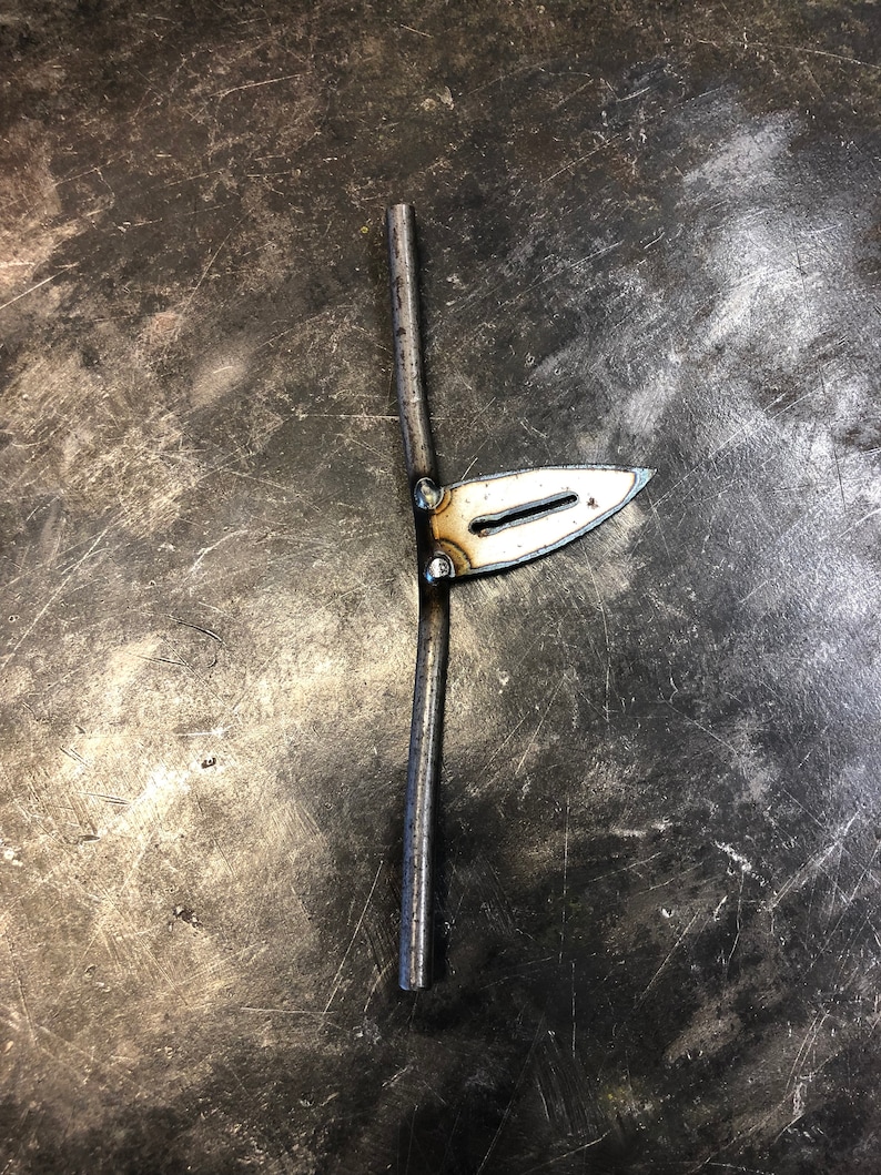 May include: A handcrafted metal tool with a blade-like shape and a vertical rod. The blade is attached to the rod with small rivets. The metal has a silver color and appears to be welded. The tool is likely used for metalworking or a similar craft.