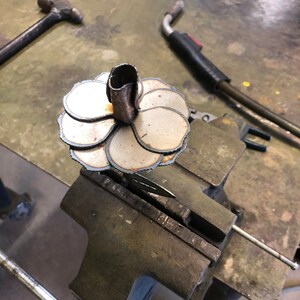 May include: A metal flower sculpture in progress, clamped in a vise. The sculpture features layered, petal-shaped pieces and a central tube. A hammer and welding torch are visible in the background, suggesting a metalworking workshop setting.