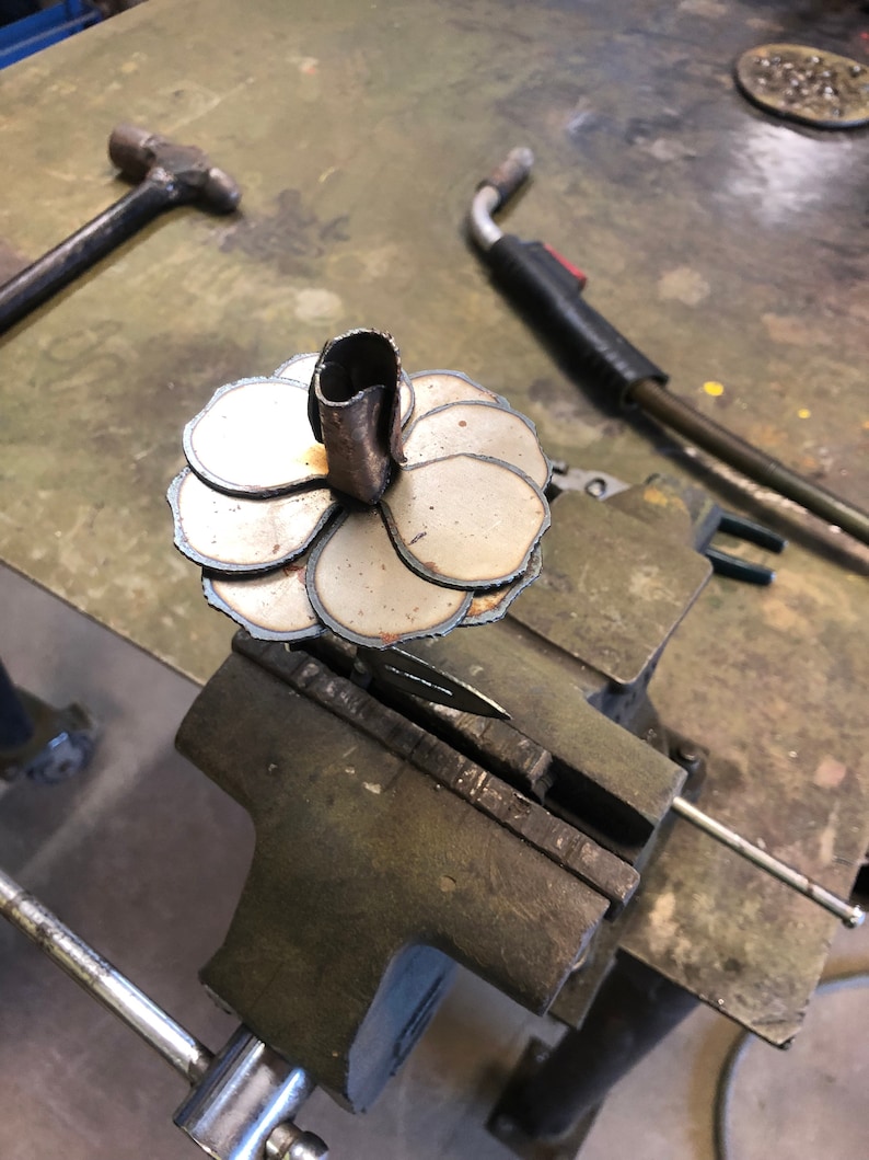 May include: A metal flower sculpture in progress, clamped in a vise. The sculpture features layered, petal-shaped pieces and a central tube. A hammer and welding torch are visible in the background, suggesting a metalworking workshop setting.
