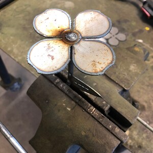 May include: A handcrafted metal flower with four petals, welded together at the center. The flower is held in a vise, showing the process of metalworking. The petals have a light brown and silver color.