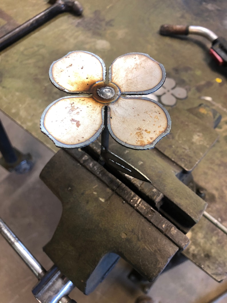 May include: A handcrafted metal flower with four petals, welded together at the center. The flower is held in a vise, showing the process of metalworking. The petals have a light brown and silver color.