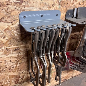 May include: A gray metal tool holder mounted on a wooden surface, holding several locking pliers. The holder is rectangular with a curved top and is secured with screws. The pliers are silver and arranged in a row.
