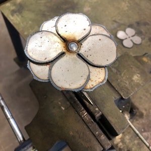 May include: A handcrafted metal flower sculpture with layered petals and a central rivet. The flower is made of silver-colored metal with visible weld marks and a rustic appearance. A smaller, similar flower shape is visible in the background.