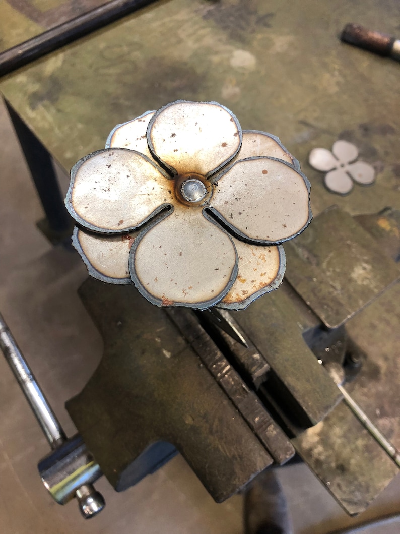 May include: A handcrafted metal flower sculpture with layered petals and a central rivet. The flower is made of silver-colored metal with visible weld marks and a rustic appearance. A smaller, similar flower shape is visible in the background.