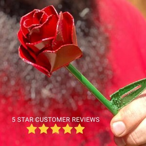 May include: A handcrafted metal rose with a vibrant red bloom and a green stem and leaf. The rose is held in a person's hand. The text "5 STAR CUSTOMER REVIEWS" is displayed below the rose.