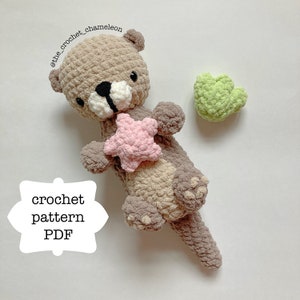 Op de afbeelding: Haakpatroon PDF voor een bruine en beige amigurumi otter die een roze gehaakte ster houdt. De otter heeft zwarte ogen en een zwarte neus. Er is een groen gehaakt hart op de achtergrond.