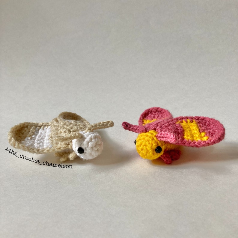 May include: Two crocheted moths, one beige with white stripes and one yellow with pink wings. The moths have black eyes and are facing the camera.