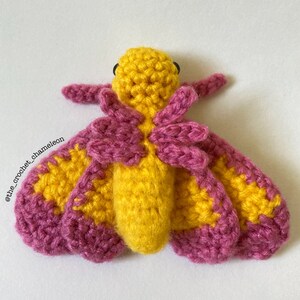 May include: A crocheted moth with yellow and pink wings. The moth has black eyes and a fuzzy body.