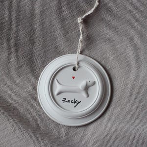 May include: A white, round ceramic tag with a dachshund dog design and the name "Rocky" in black script. A small red heart is above the dog. The tag is attached to a white string.