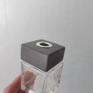 Puede incluir: Una botella de vidrio transparente con forma cuadrada y una tapa de madera gris. La tapa tiene un pequeño agujero redondo en el centro.