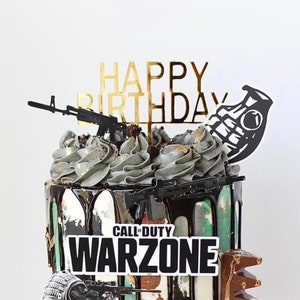 以下が含まれることがあります： 迷彩柄の誕生日ケーキで、チョコレートのドリップアイシングとコールオブデューティウォーゾーンのロゴが施されています。ケーキは、黒と金の「ハッピーバースデー」の飾り、黒いおもちゃの銃、黒い手榴弾で飾られています。