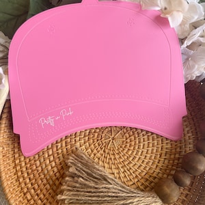 May include: Pink blank baseball cap with the text "Pretty in Pink" on the brim. The cap is sitting on a woven basket with a brown tassel.