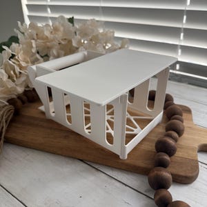 Puede incluir: Una cesta de almacenamiento blanca y rectangular con una parte superior plana y lados ranurados verticales. La cesta está sobre una tabla de cortar de madera, con una hebra de cuentas de madera marrón y flores blancas artificiales en el fondo.