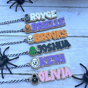 May include: Six Halloween-themed gift tags with names written in colorful, bold letters. Each tag features a different spooky character, including a ghost, a pumpkin, a skull, a monster, and a black cat.