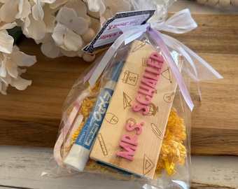 May include: A clear plastic bag filled with yellow shredded paper, a pink dry-erase marker, and a wooden sign with the text "Mrs. Schaller" in pink letters. The bag is tied with a white ribbon and a small tag that says "Thank You & Happy First Day of School!"