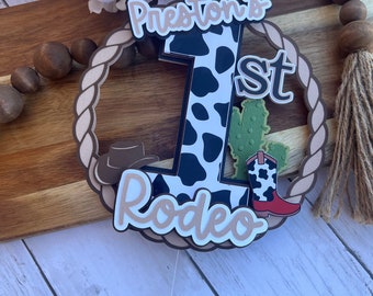 May include: A cake topper featuring a cow print number one, a cowboy hat, a cactus, and a cowboy boot. The topper is surrounded by a rope design and the words "Preston's 1st Rodeo".