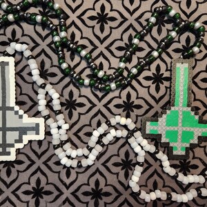 May include: Two beaded necklaces with black, white, and green beads. One necklace has a gray upside-down cross pendant, and the other has a green upside-down cross pendant.