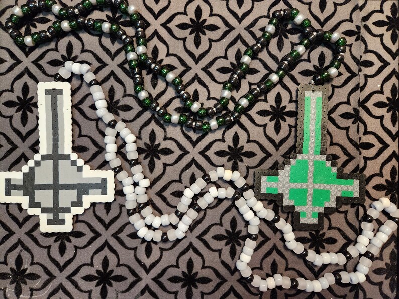 May include: Two beaded necklaces with black, white, and green beads. One necklace has a gray upside-down cross pendant, and the other has a green upside-down cross pendant.