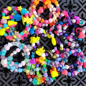 May include: A collection of colorful beaded bracelets with various charms, including stars, smiley faces, hearts, and dice. The bracelets are made of plastic and are perfect for stacking or wearing individually.