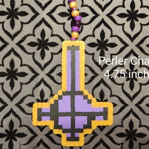 May include: Purple and black upside down cross charm made with perler beads. The charm is outlined in yellow and measures 4.75 inches.