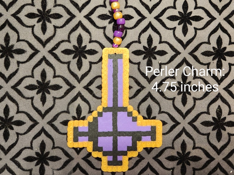 May include: Purple and black upside down cross charm made with perler beads. The charm is outlined in yellow and measures 4.75 inches.