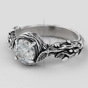 May include: A silver ring with a large, clear, round gemstone. The ring has an intricate floral design with leaves and vines around the band and gemstone setting. The ring is set against a white background.