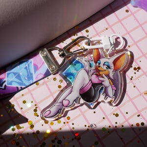 Könnte beinhalten: Ein durchsichtiger Acryl-Schlüsselanhänger zeigt eine weiße und rote Comicfigur mit Fledermausflügeln, die ein schwarzes und pinkfarbenes Outfit trägt. Die Figur ist in einer verführerischen Pose dargestellt, mit einem hochgehobenen Bein und einem herzförmigen Objekt in der Hand. Der Schlüsselanhänger ist an einem bunten Band mit einem silbernen Verschluss befestigt.