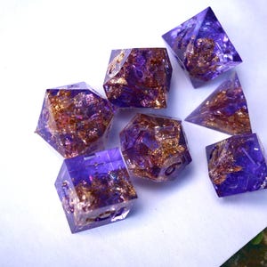 May include: Set of seven polyhedral dice in various shapes. The dice are a translucent purple with gold flakes suspended inside. The dice are arranged on a white surface.