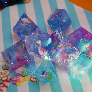Puede incluir: Un conjunto de dados iridiscentes con varios colores y diseños, incluyendo un flamenco, una sandía y un arcoíris. Los dados están hechos de resina transparente y tienen un acabado brillante.