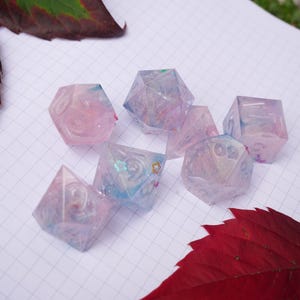 May include: A collection of translucent, multi-sided dice in shades of pink, blue, and purple. The dice are scattered on a white grid, with red and green leaves in the background. These dice are perfect for tabletop games.