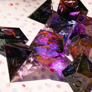 May include: A collection of multi-sided dice in various shapes, including pyramids and multi-faceted forms. The dice are translucent with a mix of colors, including purple, orange, and green. The dice are scattered on a white surface with colorful polka dots.