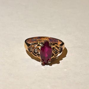 May include: A gold ring with a central, marquise-cut ruby. The ring has a decorative band with small, clear accent stones. The ruby is a deep red colour, and the ring is set against a neutral background.