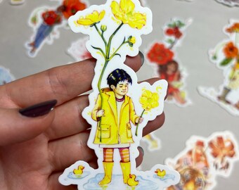 May include: A sticker depicting a person in a yellow raincoat and wellington boots holding yellow flowers. The person stands in a puddle with rubber ducks. The sticker has a white border on a light grey background.