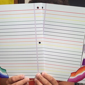 以下が含まれることがあります： 虹色の線と、さまざまなプライドフラッグのデザインが施された装飾的な縁取りが施された白いノート。 ノートはスパイラル綴じで、バインダーに挿入するための3つの穴があります。 ノートは開いており、罫線入りのページが表示されています。