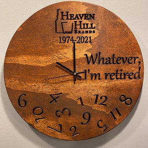May include: A round wooden clock with a dark brown finish. The clock face features the text "Heaven Hill Brands 1974-2021" and "Whatever, I'm retired." Black hands indicate the time. Numbers are carved into the wood.