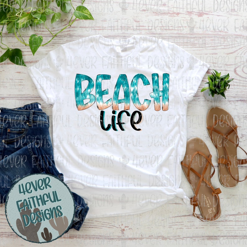May include: White t-shirt with a teal and orange "Beach Life" graphic. The shirt is laid out on a wooden surface with a pair of brown sandals and a potted plant.