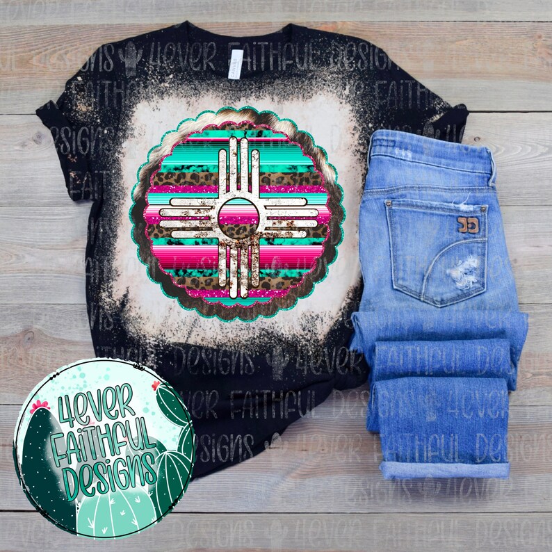 Peut inclure: Un t-shirt noir avec un motif blanchi repr&eacute;sentant un symbole du Sud-Ouest turquoise, rose et marron avec des accents de paillettes. Le symbole est entour&eacute; d'un cercle blanc. Le t-shirt est sur une surface en bois avec un jean bleu.