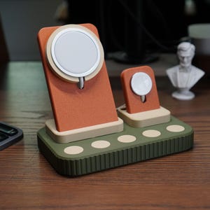 May include: A multi-device charging station in shades of orange, green, and beige. The station has two charging pads, one for a phone and one for a watch. The base is green with beige circles. The charging station is on a wooden surface.