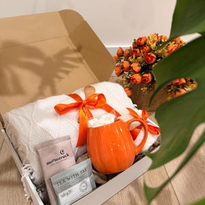 May include: A gift box containing a white cable-knit blanket tied with an orange ribbon, a bright orange pumpkin-shaped ceramic mug filled with white marshmallows, a Moonstruck chocolate bar, and a Tea With Tae tea bag, all nestled inside.