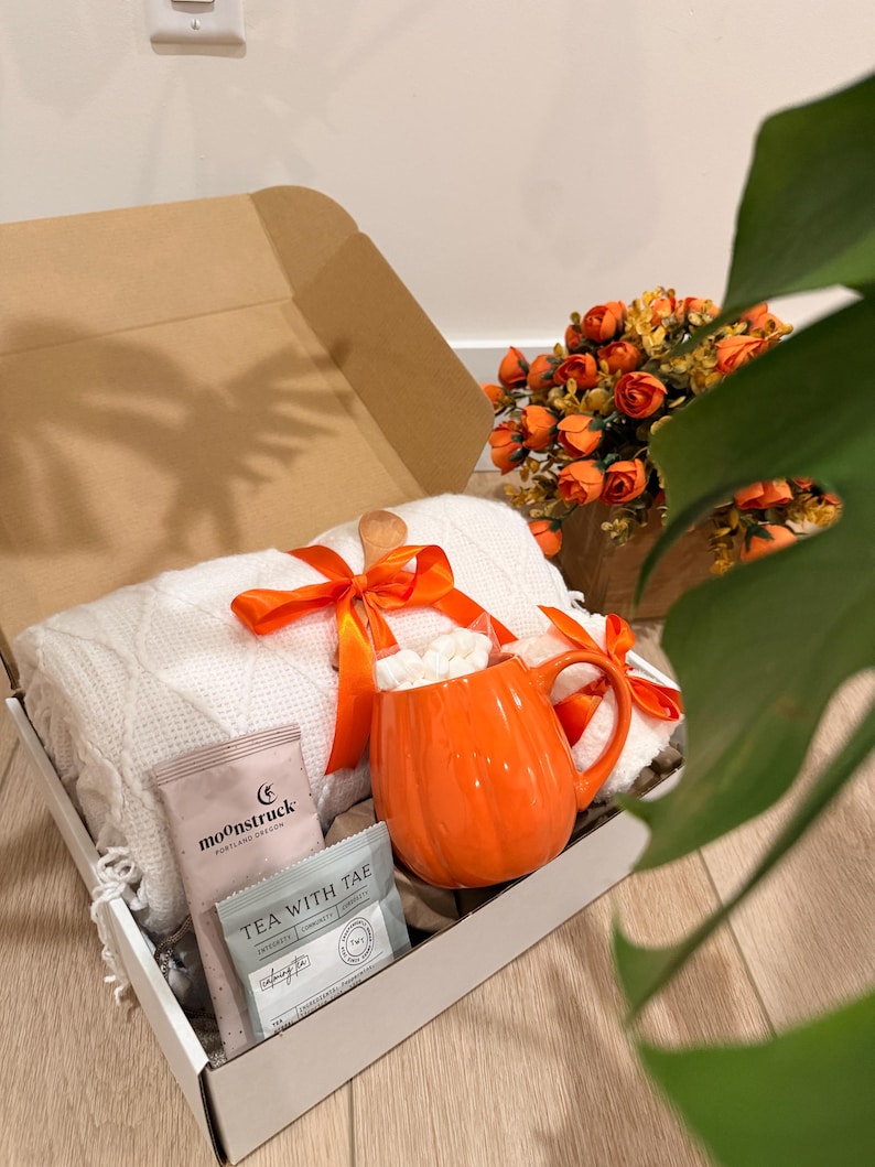 May include: A gift box containing a white cable-knit blanket tied with an orange ribbon, a bright orange pumpkin-shaped ceramic mug filled with white marshmallows, a Moonstruck chocolate bar, and a Tea With Tae tea bag, all nestled inside.