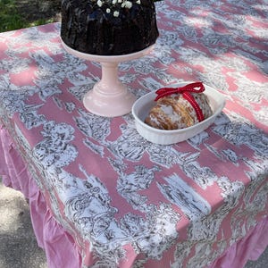 May include: A pink tablecloth with a white floral pattern and ruffled edges. A chocolate cake with chocolate frosting and strawberries on top is on a white cake stand. A glazed pastry is in a white oval dish with a red ribbon.