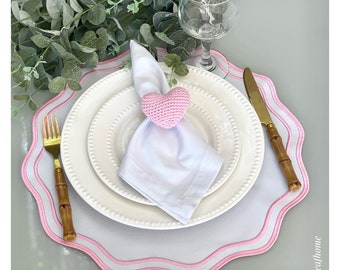 May include: White dinner plate with a pink crocheted heart napkin ring on a white napkin. The plate is on a white and pink scalloped placemat.